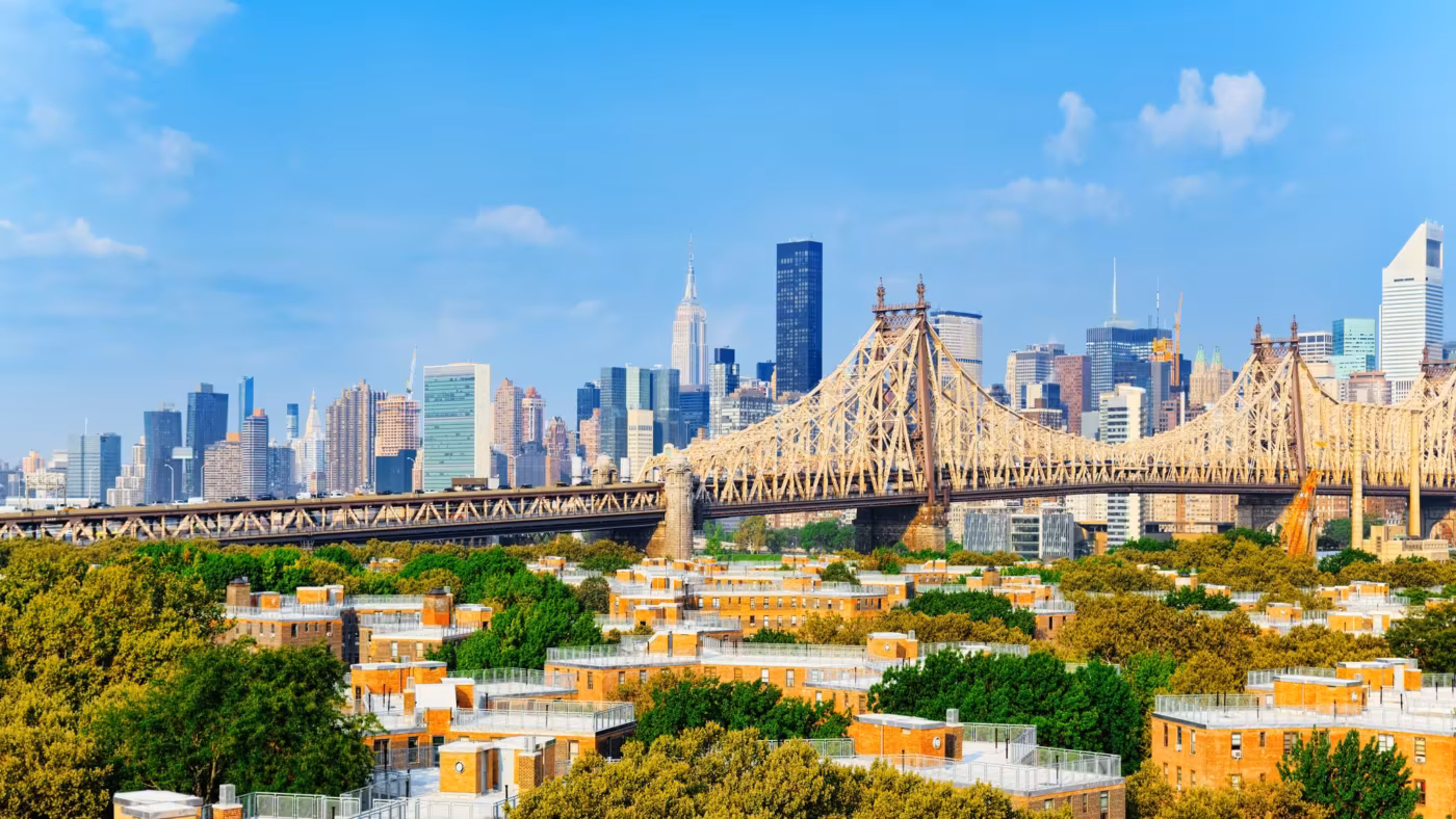 Rows of low houses and trees with the Queensboro Bridge and the Manhattan skyline behind