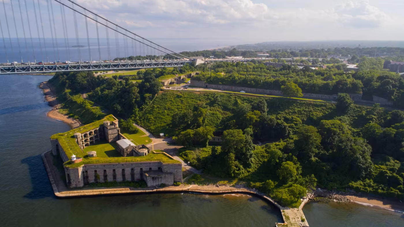 A green harbour shoreline in New York with an old fort and a suspension bridge beyond