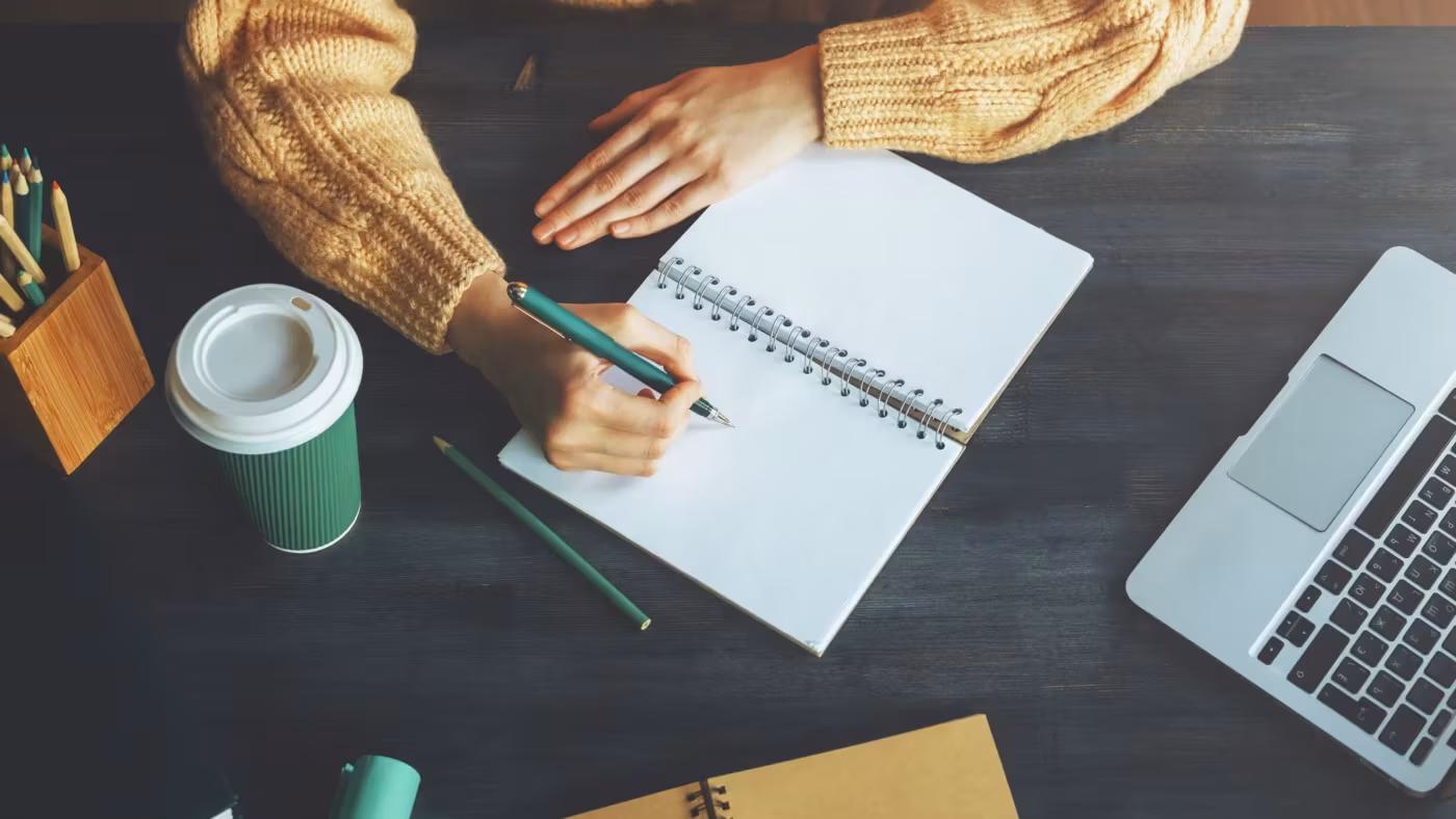A hand writing in a notebook at a desk beside a laptop
