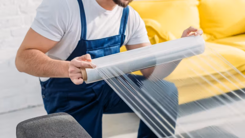 A mover in bib overalls drawing a roll of stretch film across an upholstered sofa
