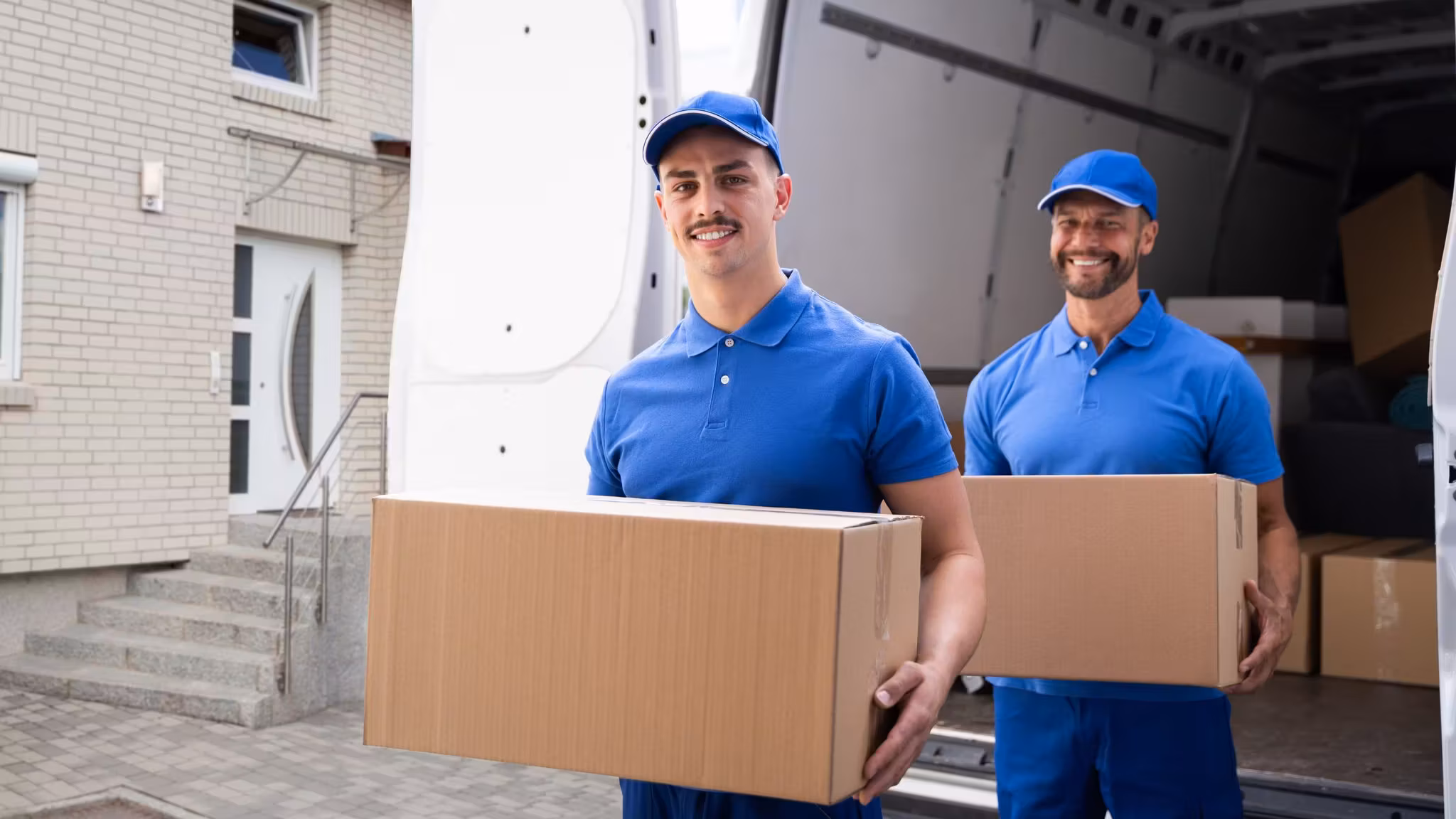Two movers with a carton at the open side of a van outside a building