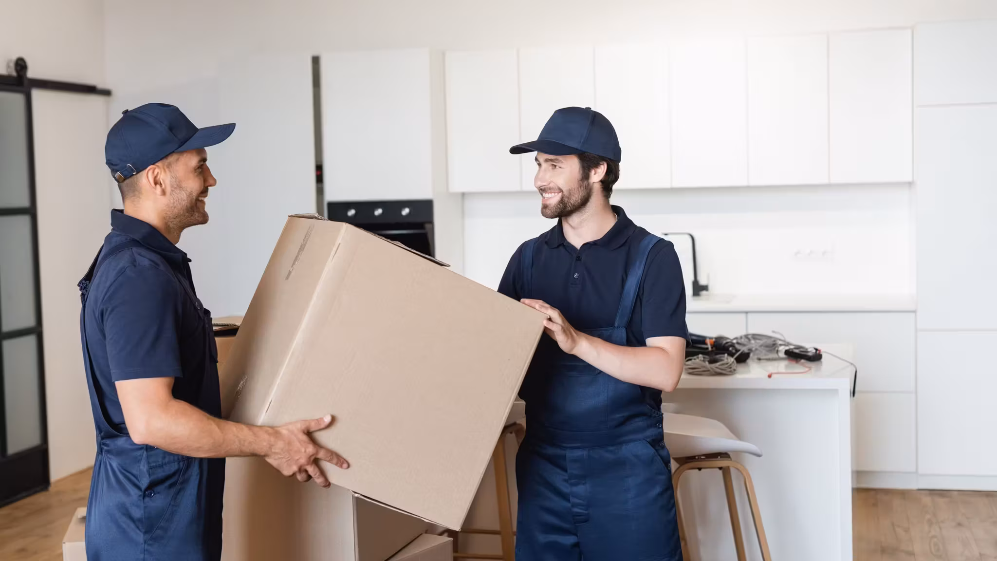 Two movers carrying a large carton between them across a kitchen