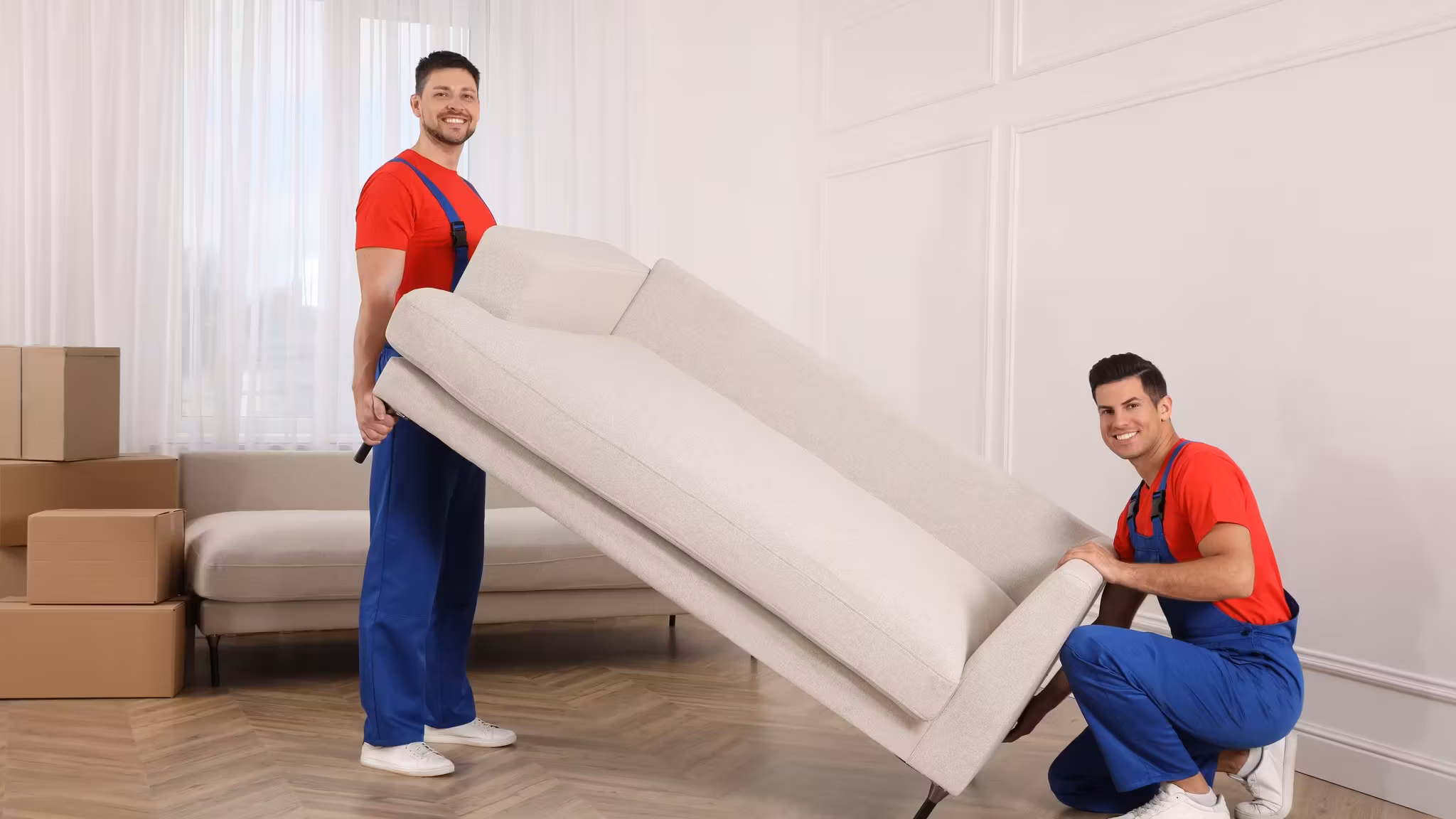Two movers setting down a cream sofa in a room with cartons behind