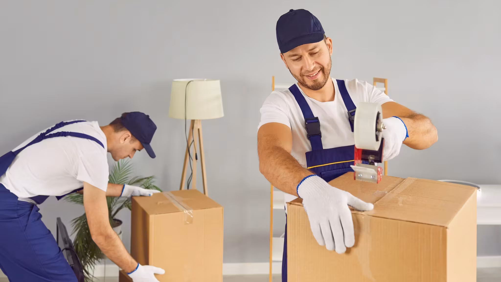 Two movers working on a taped carton in a room, one running the tape gun