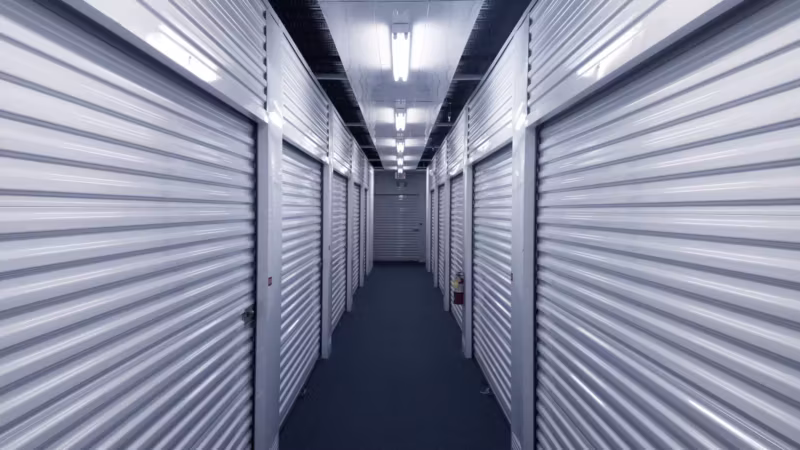 A long lit corridor between two runs of grey roller doors inside a storage building