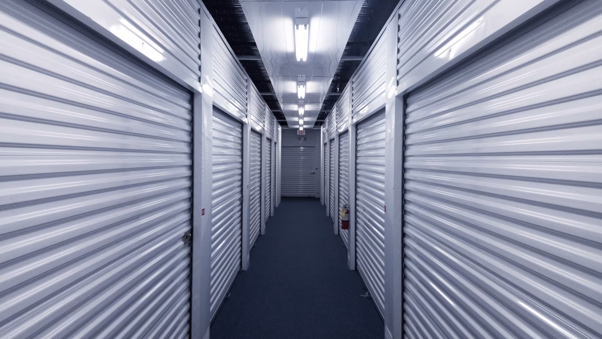 A long lit corridor between two runs of grey roller doors inside a storage building
