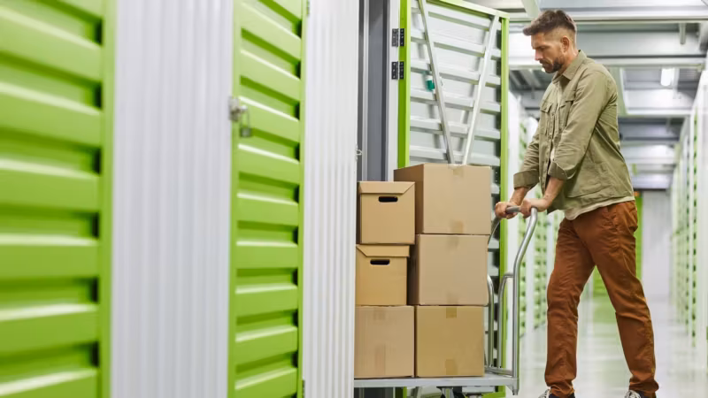 A man pushing a flatbed trolley loaded with cartons along an aisle of green unit doors