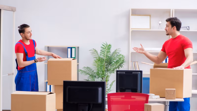 Two movers boxing a monitor and desk electronics beside a part-packed shelf unit
