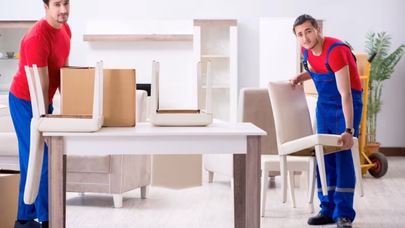 A mover setting a dining table upright in a bright room with chairs stacked beside it