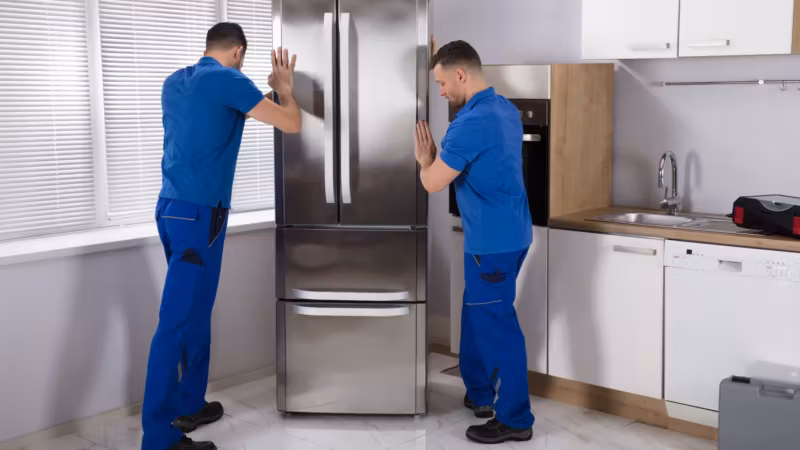 Two movers easing a refrigerator away from a kitchen wall