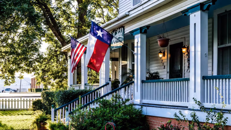 The porch of a white clapboard Texas house with the state and national flags flying