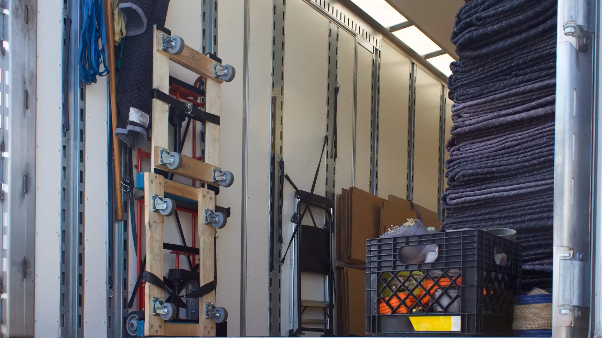 The inside of a moving trailer: folded moving blankets stacked high, hand trucks racked against the wall and straps hanging ready
