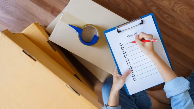 A hand ticking items on a printed checklist held over an open carton