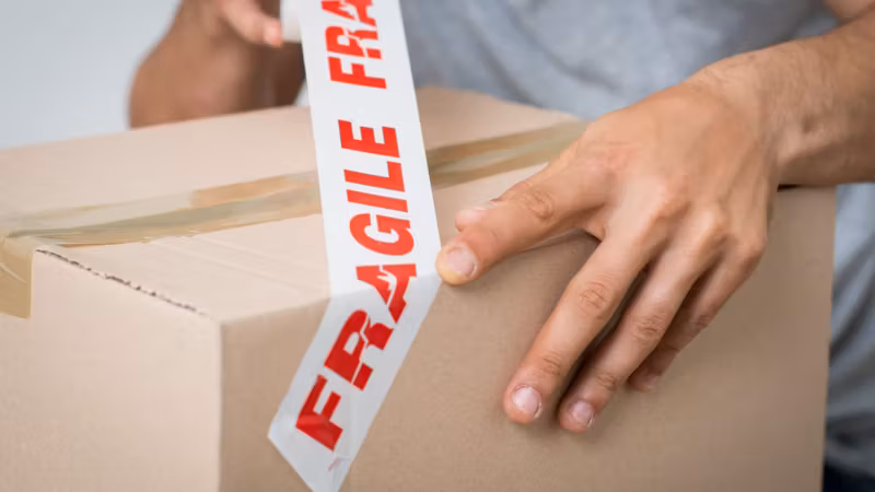 A hand running red FRAGILE tape across the seam of a carton