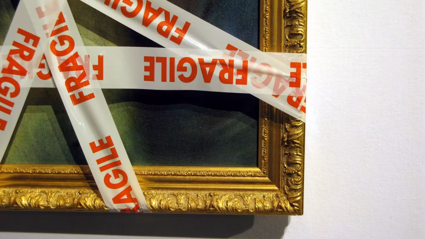 A gilt-framed picture criss-crossed with red FRAGILE tape, propped against a wall