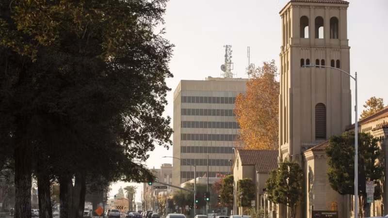 A tree-lined street of low offices with a bell tower behind