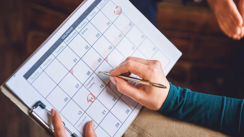 A hand marking a date on a wall calendar with a pen