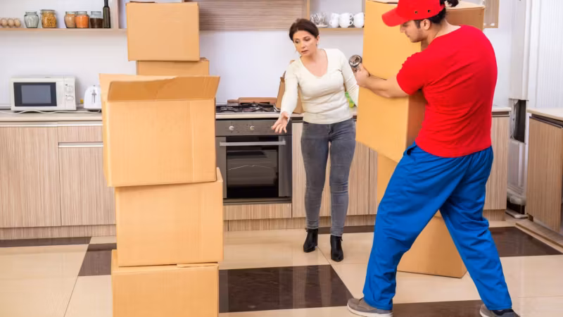 A mover carrying a carton through a kitchen as a resident stands aside