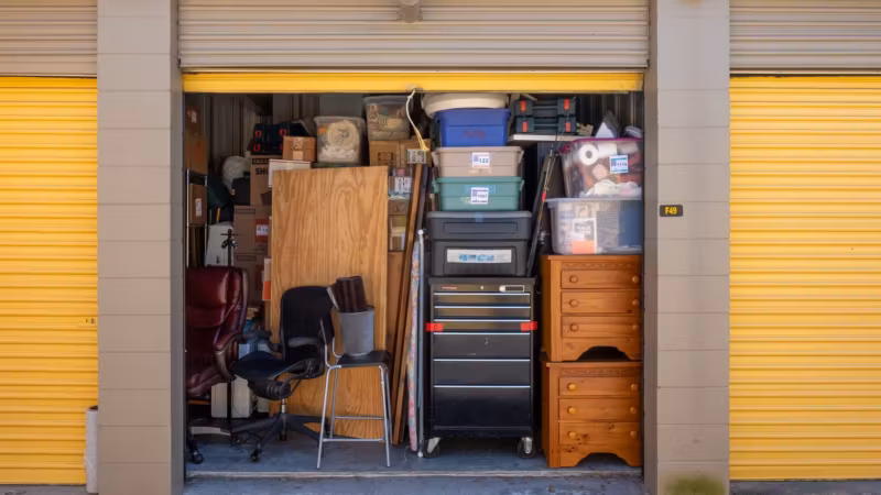 A storage unit packed with a chest of drawers, crates and stacked boxes behind a raised door